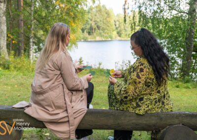 Kaksi naista istuu järven rannalla lehtiä käsissään.