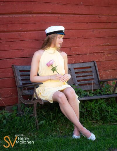Ylioppilas istuu penkillä punaisen seinän edessä ja pitelee kukkia.